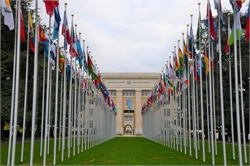 [ai] A view of the United Nations building in Geneva, framed by rows of international flags on tall poles, set against a cloudy sky.